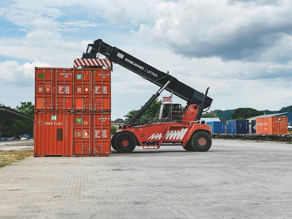 Red industrial crane lifting stacked cargo containers at a container yard in Ipoh, Malaysia.